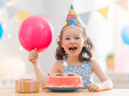 Dziecko siedzi w urodzinowej czapce z tortem i balonem w swoim domu.