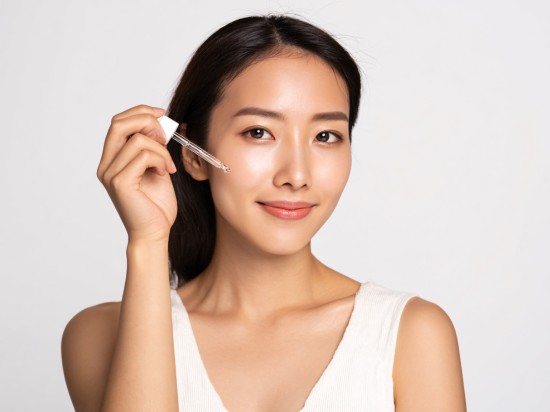 Uśmiechnięta kobieta nakłada szklaną pipetę na policzek serum do twarzy jako element codziennej pielęgnacji.