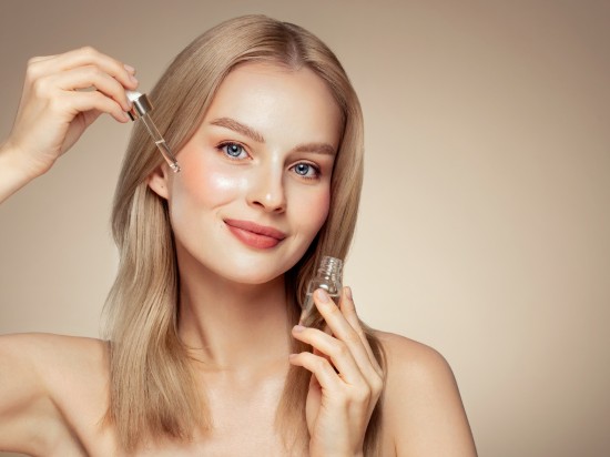 Uśmiechnięta kobieta nakłada na policzek szklaną pipetą serum do twarzy.