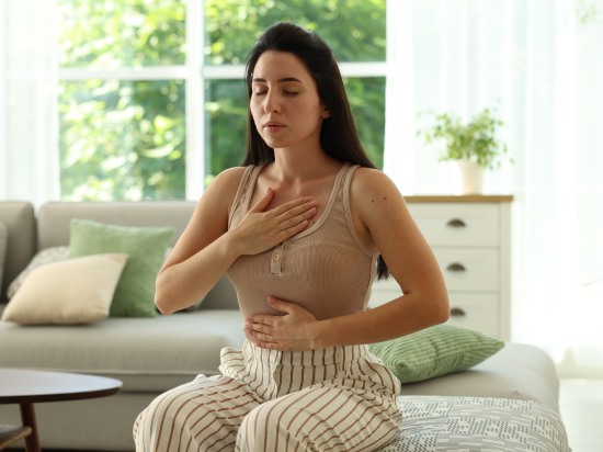 Kobieta z zamknietymi oczami siedzi na łóżku i ćwiczy oddech aby obniżyć poziom kortyzolu o poranku.