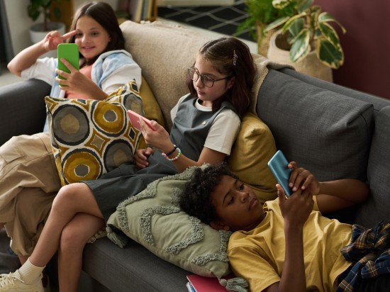 Trójka znudzonych dzieci leży na kanapie i wpatruje się w swoje telefony, co nie jest dla nich zdrowym nawykiem.