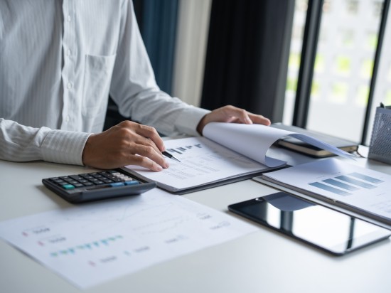 Osoba siedzi przy biurku z dokumentami i kalkulatorem i robi rozliczenia finansowe.